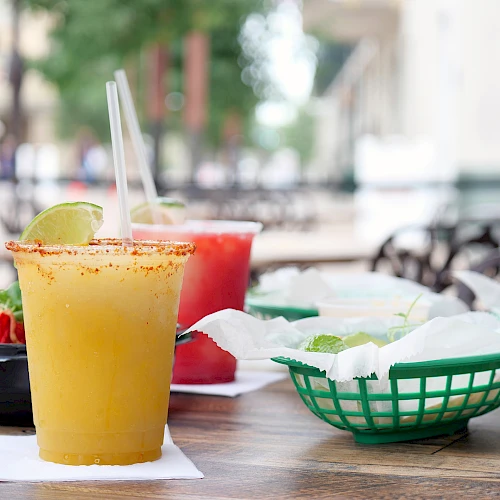 A sunny outdoor meal: a mango or orange smoothie with a spice rim, straw, lime wedge, plus bowls of food and chips on a communal table outdoors.