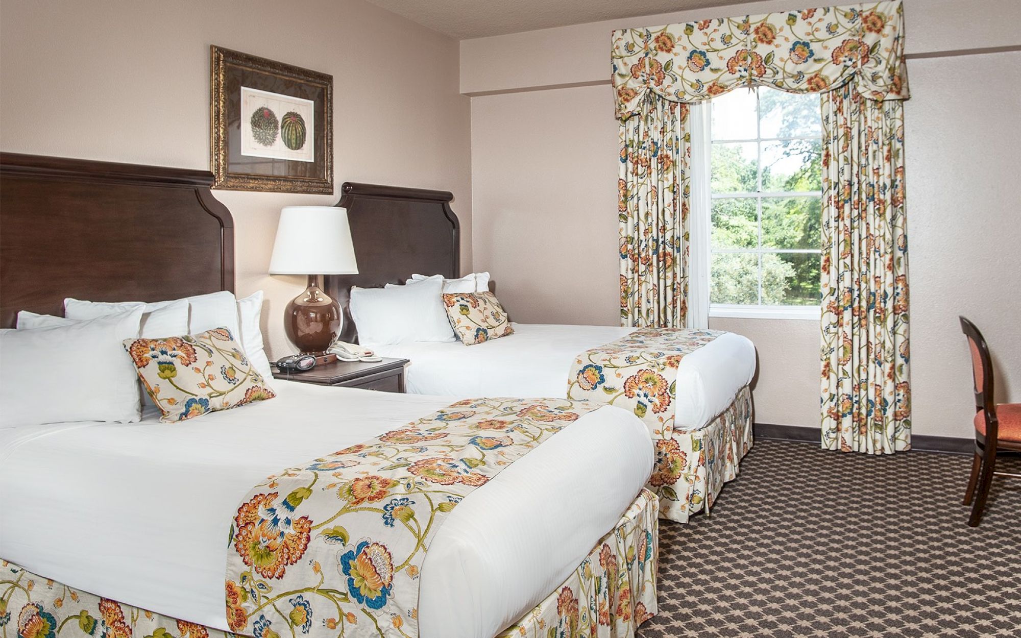 A hotel room with two beds, floral bedding, patterned curtains, a nightstand, lamp, wall art, and a window with outside view.