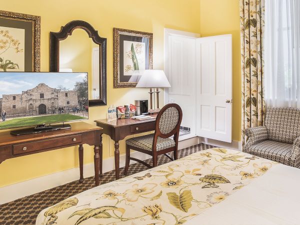 A cozy hotel room with a patterned bedspread, TV, desk, chair, and lamp. Yellow walls feature framed art and a large window with curtains.