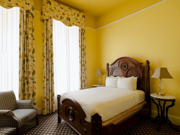 A yellow-walled hotel room with a carved wooden bed, white bedding, floral drapes, a seating chair, and a side table with a lamp.