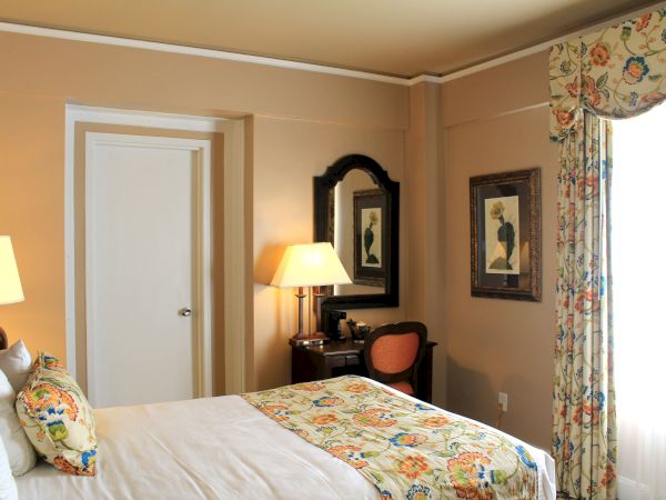 A cozy hotel room with a bed, floral bed runner and pillows, two lamps, a dresser with a mirror, a door, framed art, and a large window with floral curtains.