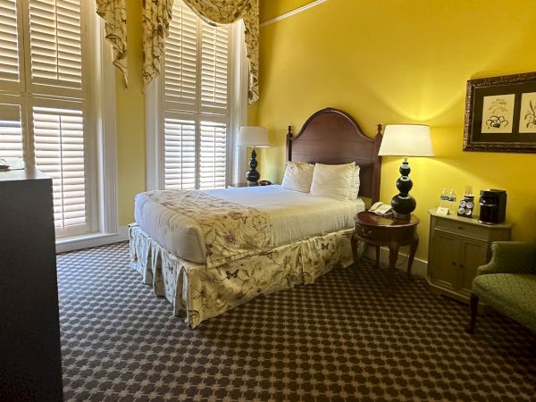A cozy hotel room with yellow walls, a bed, armchair, and two lamps. There's a TV, artwork, and a window with shutters.