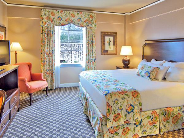A cozy hotel room with a large bed, decorative pillows, a TV, an armchair, lamps, and floral curtains and bedspread.