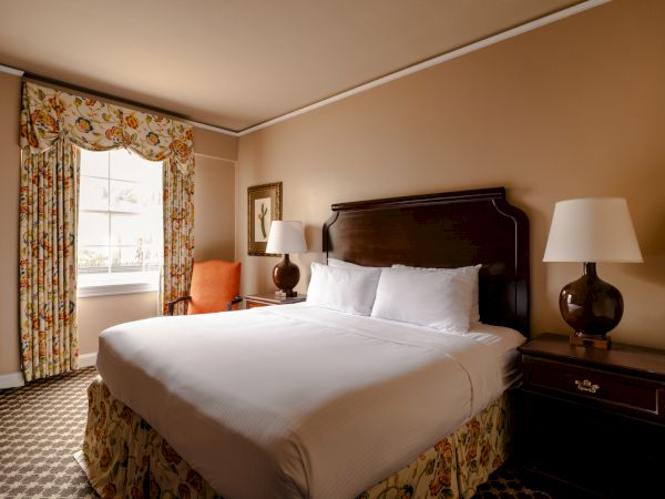 A hotel bedroom with a king bed, white linens, dark wood headboard and nightstands, two lamps, an orange chair, floral drapes, and a window light.