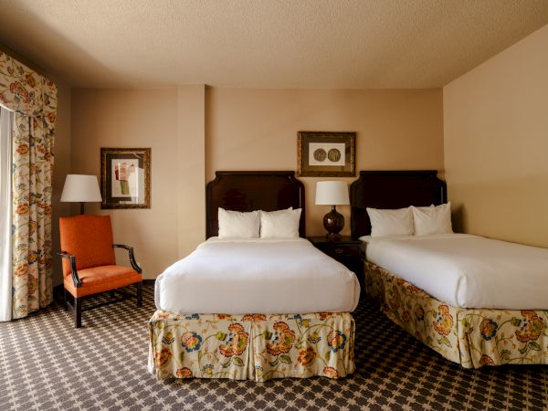 Two white‑duvet hotel beds with floral bedskirts, a nightstand between them, a lamp, a framed picture on the wall, and a seating chair with a lamp in a patterned carpeted room.
