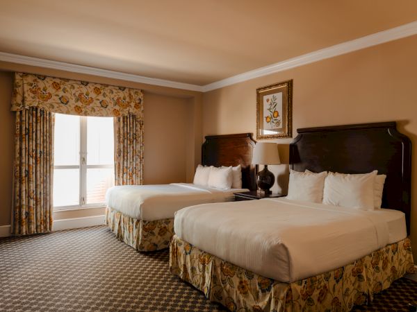 Two double beds in a beige-toned hotel room with floral curtains, a window, and framed artwork above the headboards. 140 characters, ending with a period.