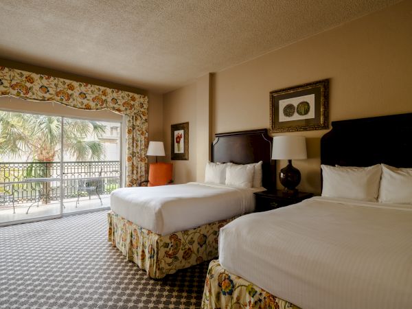 Two hotel beds with floral skirts, a balcony view of palm trees, lamps, and framed art on the wall.