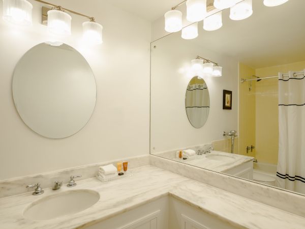 A bright bathroom with marble countertops, double sinks, round mirrors, and a large vanity light fixture above the mirror.