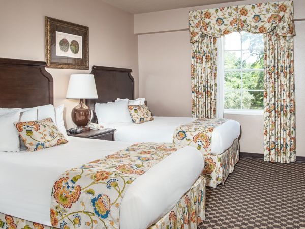 The image shows a hotel room with two beds, floral-patterned bedding, a lamp, a window with matching curtains, and a framed picture.
