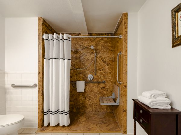 A compact bathroom with a wood-paneled shower area, white shower curtain, grab bars, a small shelf, and a dark wood vanity with stacked towels.