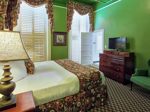 A green-walled bedroom with a bed, patterned bedding, armchair, lamp, dresser, TV, and two tall windows with floral curtains and shutters.