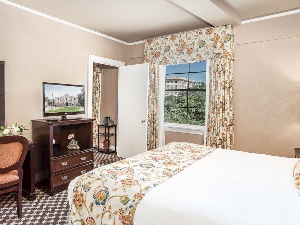A hotel room features a bed, TV, desk with chair, floral curtains, and patterned carpet, creating a cozy and inviting atmosphere.