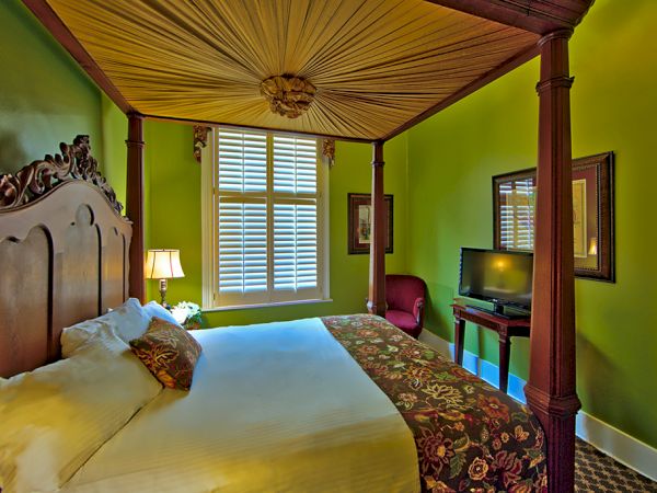 A room with a four-poster bed, green walls, a TV, and a window with shutters, featuring warm lighting and decorative elements.