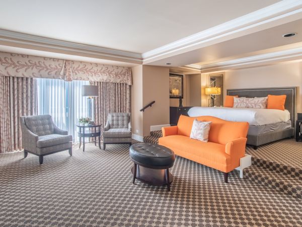 This image shows a spacious hotel room with a bed, orange sofa, chairs, and a carpeted floor.