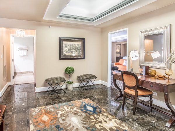 This image shows an elegantly decorated interior with a rug, desk, chairs, artwork on the wall, and a doorway leading to another room.