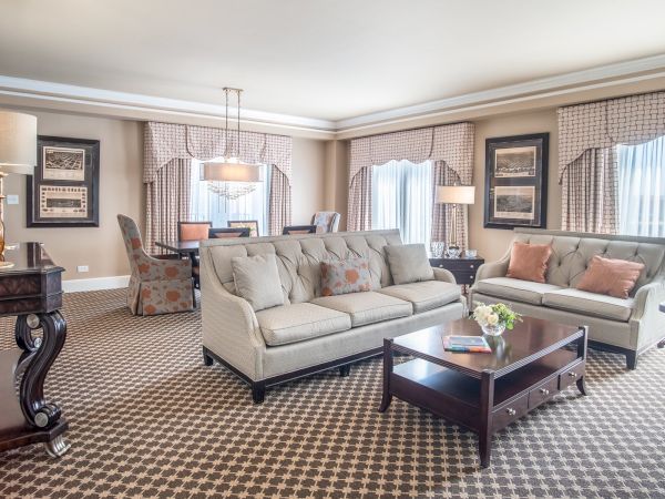 A spacious, elegant living room with sofas, lamps, artwork, and a dining area in a classic style.