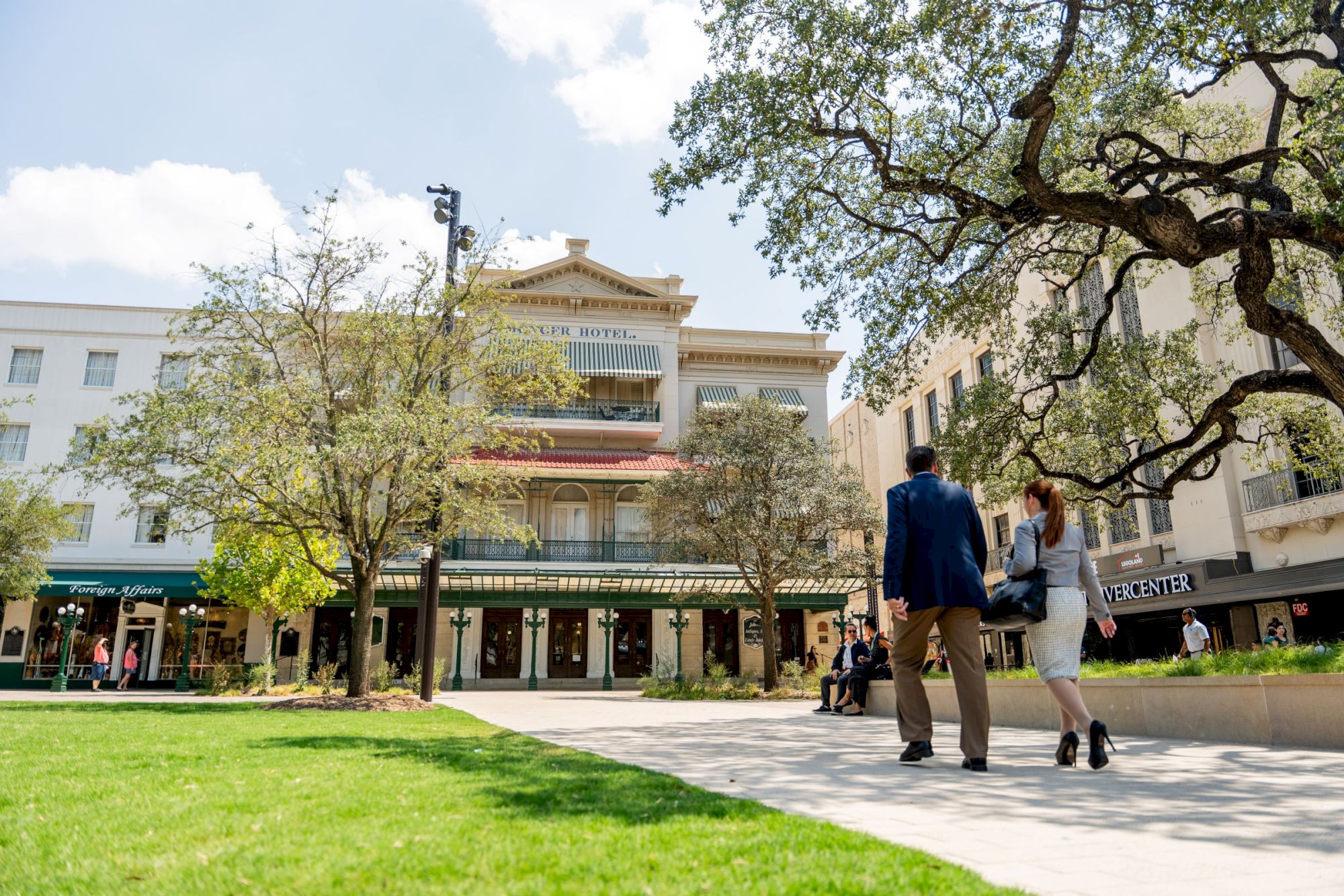 People walk on a sunny campus plaza in front of a multi-story building with trees, green lawn, and shops.