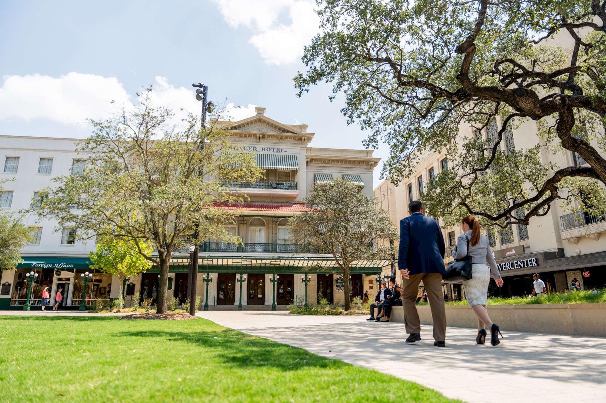 A sunny campus scene with people walking near a tree-lined plaza and a central building that resembles a university or mall.