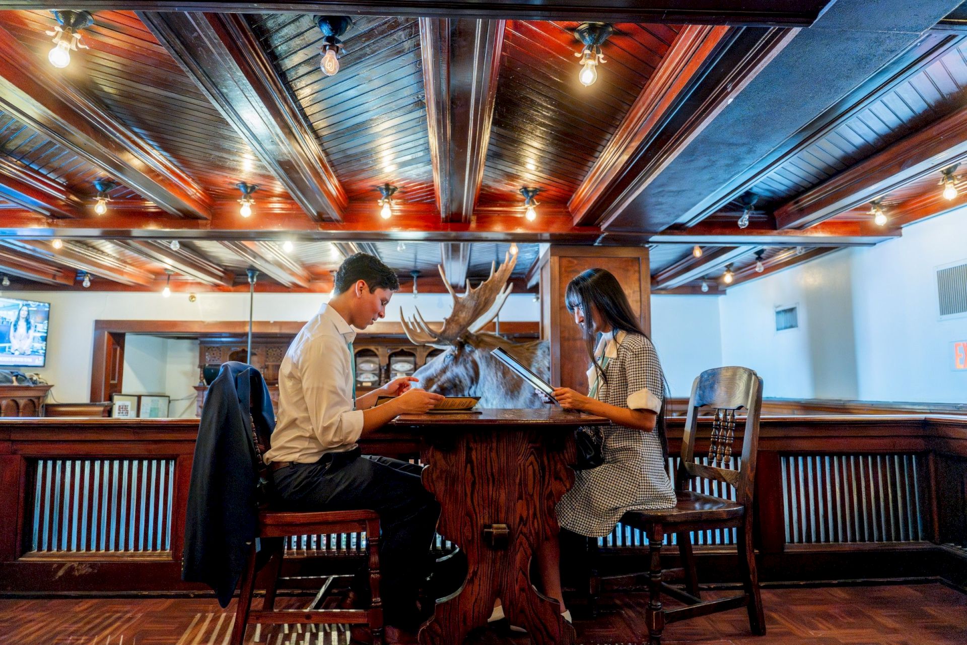 Two people sit across from each other at a rustic bar, reading menus, with a large deer head mounted on the counter between them, in a cozy, dimly lit tavern.