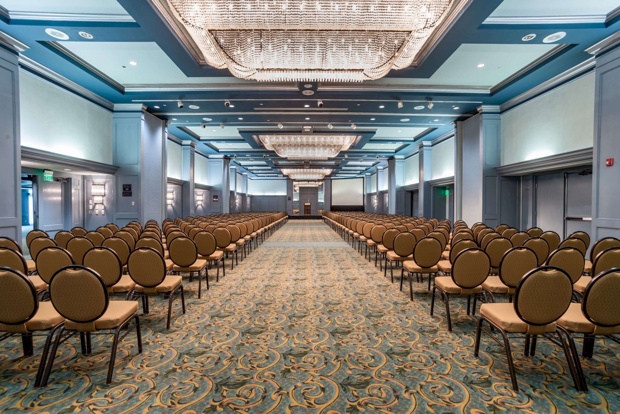 A large conference hall with rows of brown chairs facing a stage, ornate chandeliers, and carpeted floors in teal and gold tones.