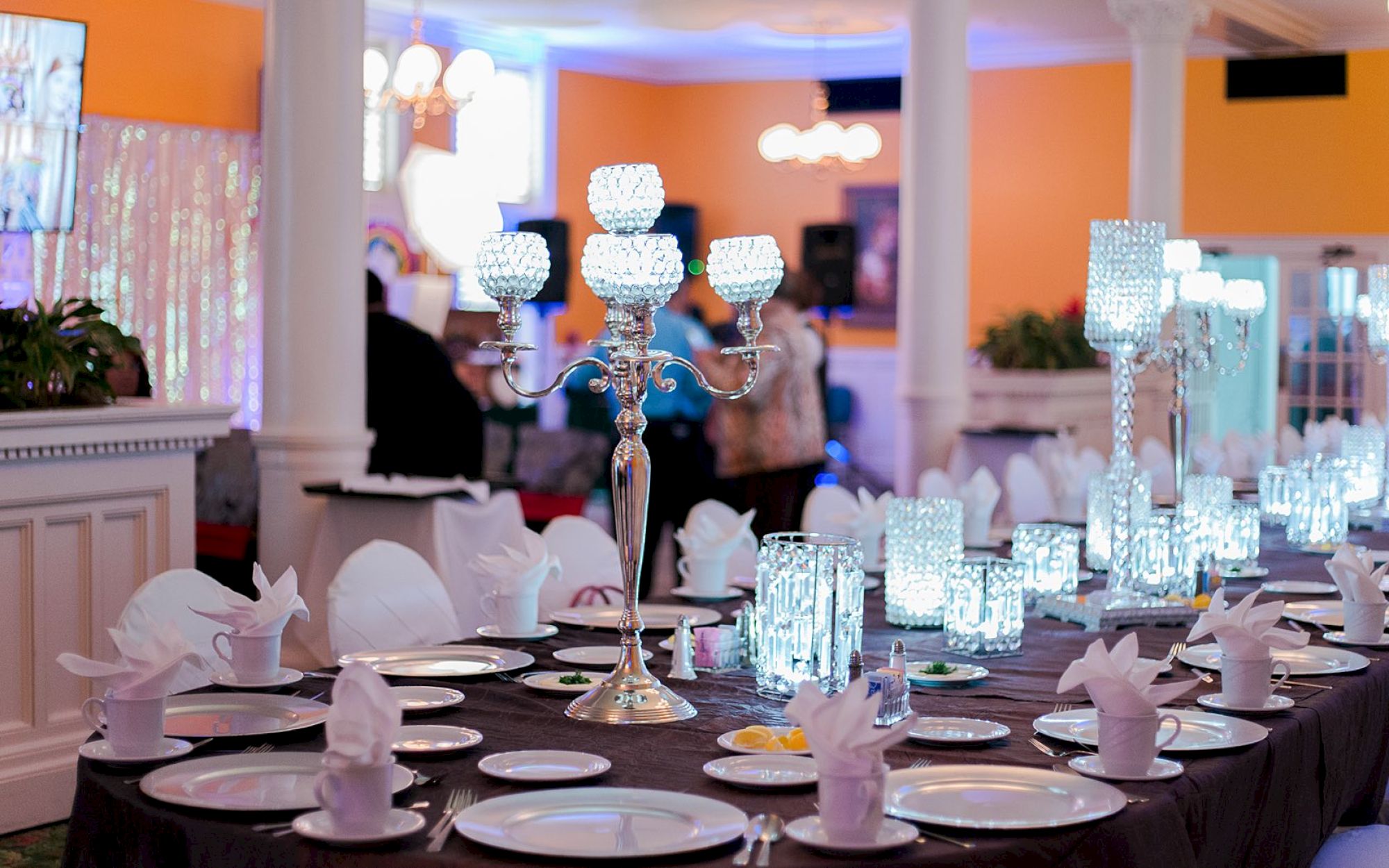 A formal dining setup with a long, dark table, white plates and napkins folded like flowers, and sparkling crystal candleholders lining the center. The background shows an orange wall and guests mingling in a softly lit room.
