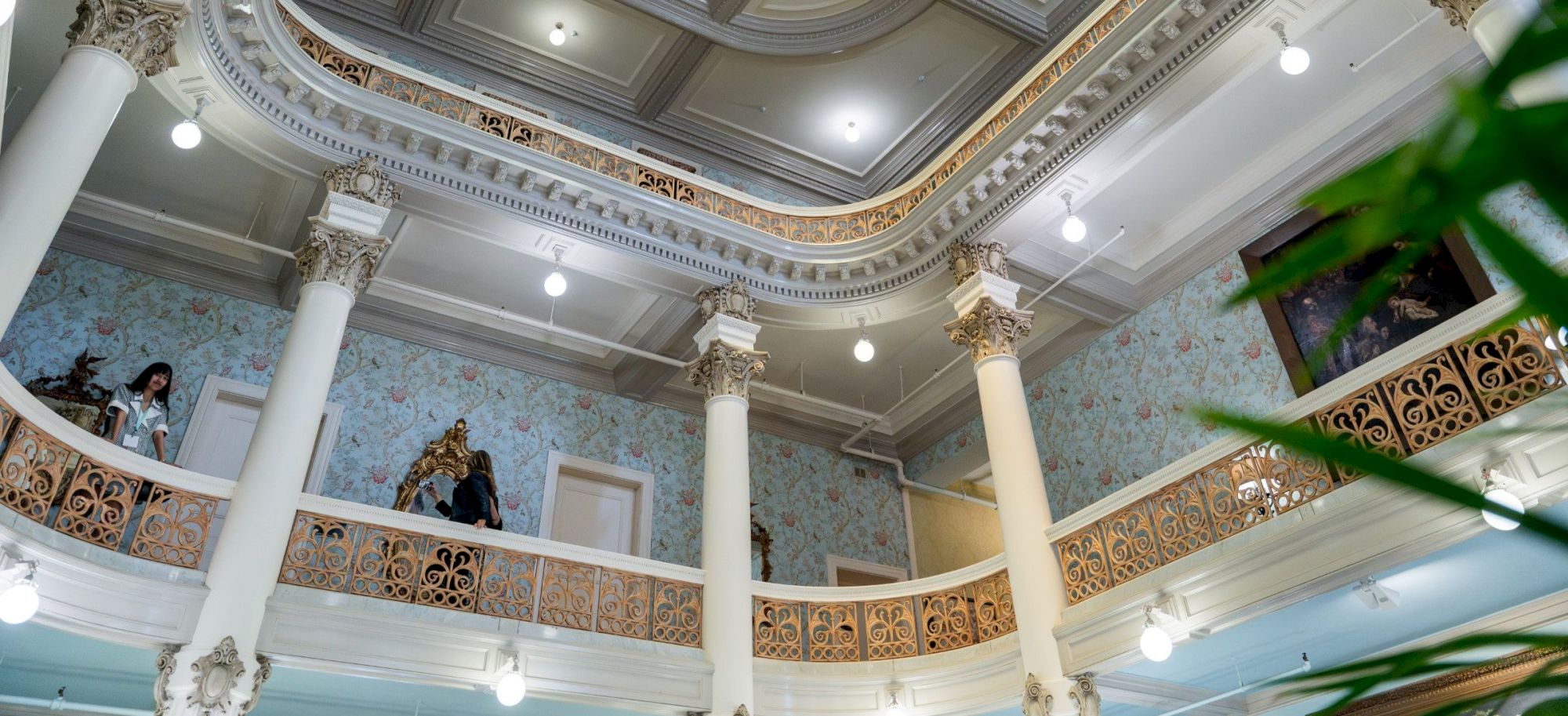 Elegant interior with ornate columns, decorative railings, and a high, patterned ceiling. Two people stand on the balcony above, ornate chandeliers hang, lush greenery in the foreground.