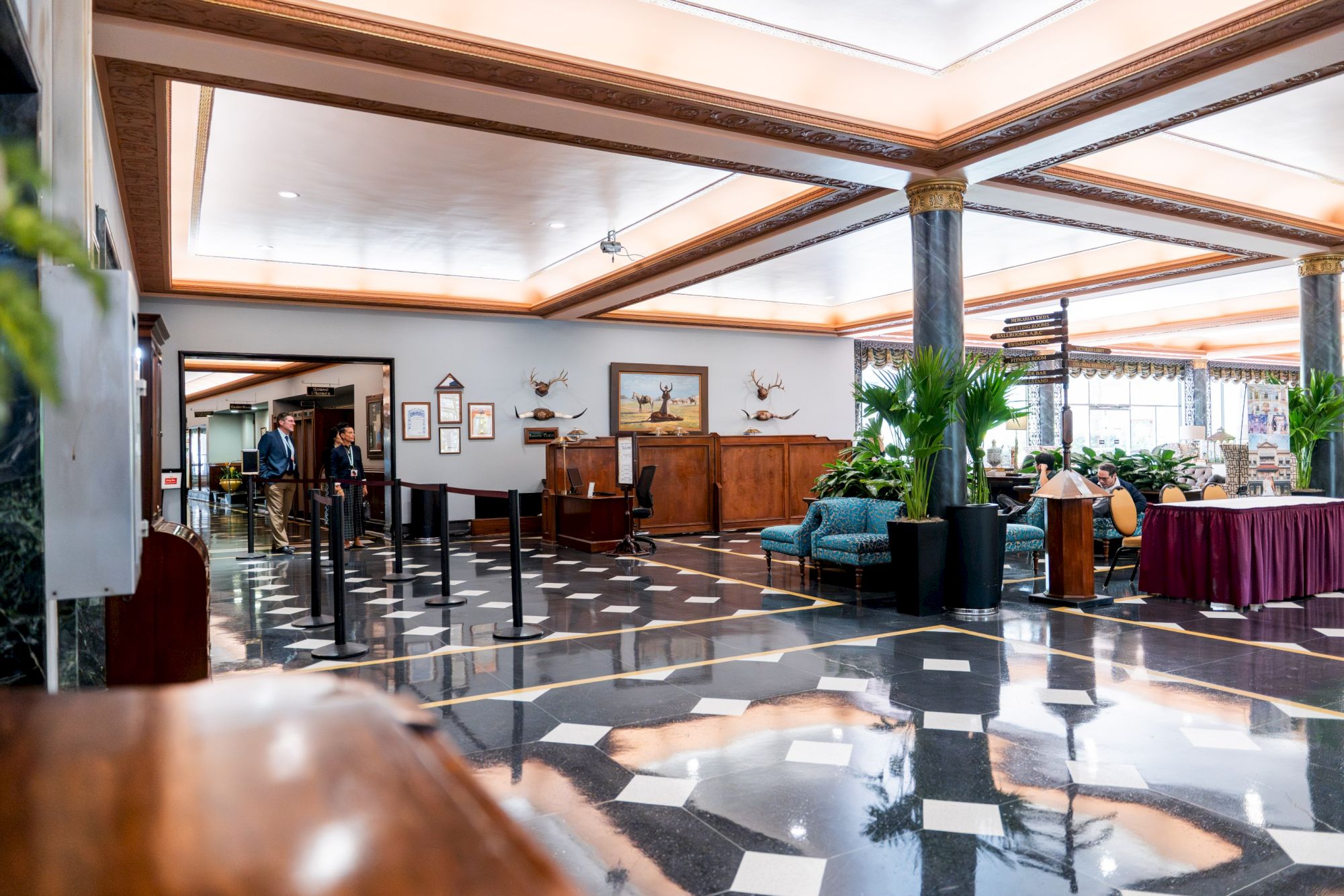 A hotel lobby with marble checkered floor, wooden beams, plants, seating areas, a reception desk, and a few guests near the back, bright and airy.