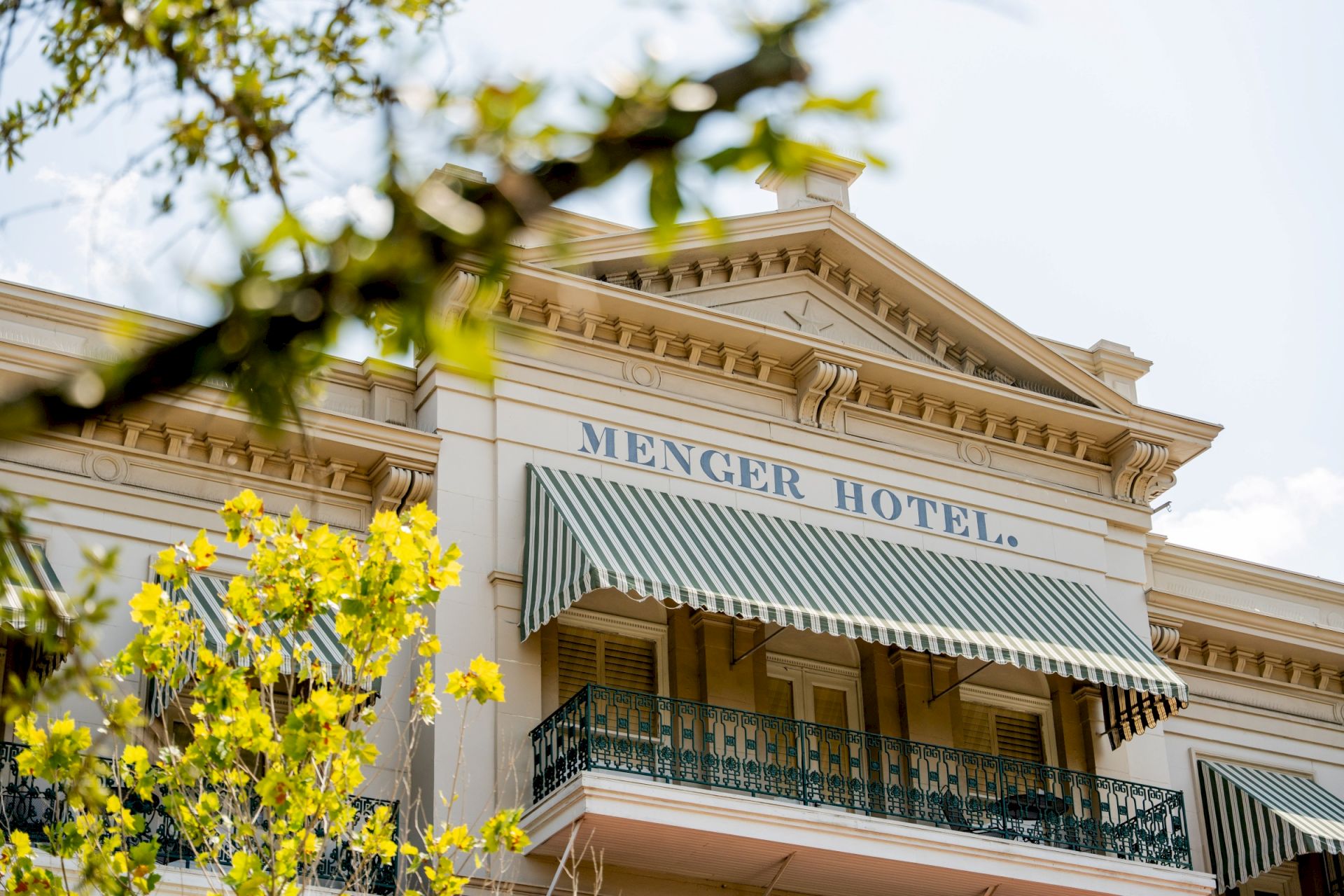 A hotel balcony and fa&ccedil;ade with a striped awning reading &ldquo;Menger Hotel,&rdquo; seen through yellow flowers and green leaves above.