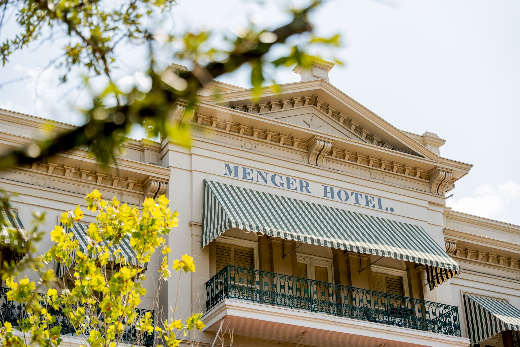 A historic hotel with a striped awning reading &ldquo;MIENGER HOTEL&rdquo; and yellow blossoms in the foreground, colonial-style railing, bright daylight.