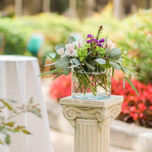A garden scene with a round white-draped table and a floral arrangement on a stone pedestal, surrounded by colorful blooms and greenery.