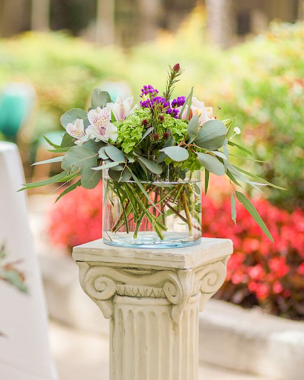A garden scene with a round white-draped table and a floral arrangement on a stone pedestal, surrounded by colorful blooms and greenery.