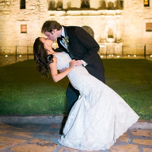 A newlywed couple shares a kiss outside a lit castle at night, the groom in a suit and the bride in a white gown, romantic and joyful.