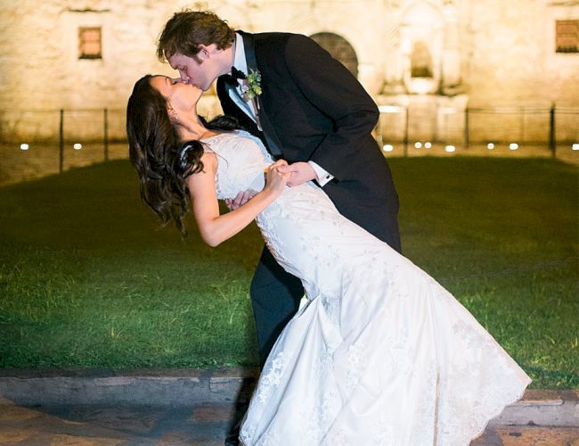 A newlywed couple shares a kiss outside a lit castle at night, the groom in a suit and the bride in a white gown, romantic and joyful.