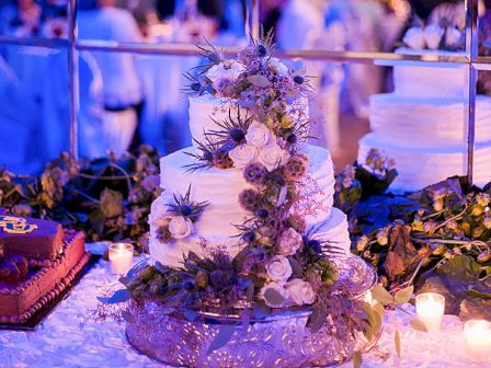 A multi-tier wedding cake decorated with white flowers and greenery, surrounded by candles and a festive backdrop.