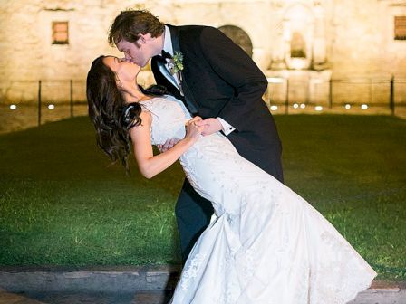 A newlywed couple shares a dramatic dip and kiss outside a lit castle backdrop at night.