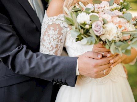 A groom in a dark suit holds a bride in a lace wedding dress, showing a bouquet of pastel roses and greenery.