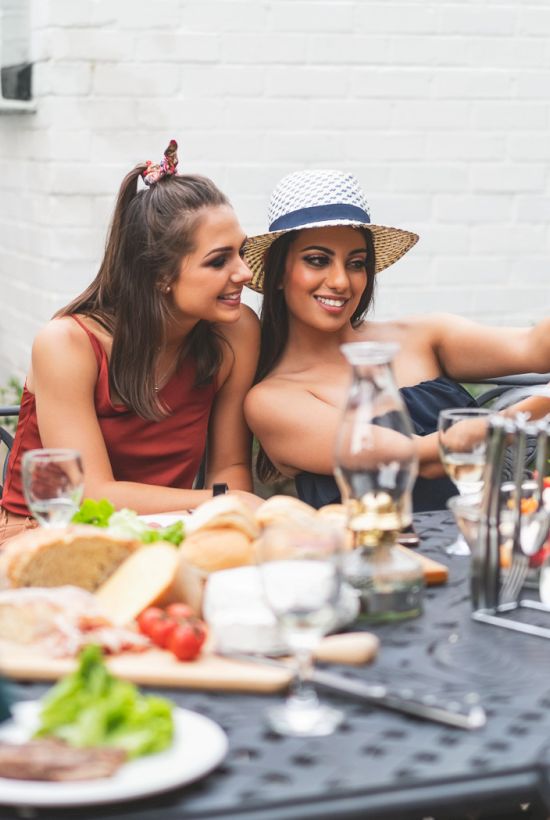 Two women laugh and toast at an outdoor table with food and drinks, enjoying a sunny meal with friends.
