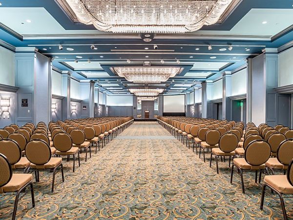 A large conference ballroom with rows of beige chairs facing a stage, ornate chandeliers, blue walls, and patterned carpet, set for a formal event.