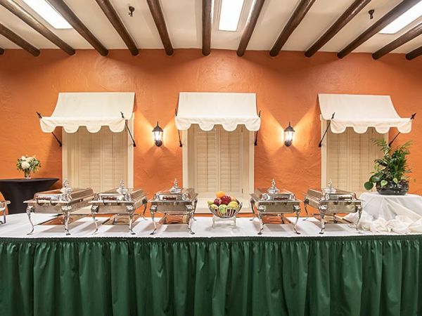 A buffet setup with chafing dishes, trays, and flowers against an orange wall with white awnings over three serving stations, skirted in green.