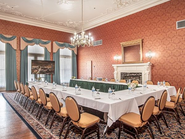 A formal, elegant conference room with a long U-shaped table, gold chairs, red wallpaper, chandelier, and a classic fireplace in a historic setting.