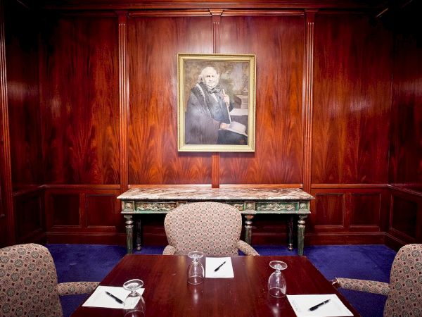 A formal dining or meeting room with dark wood-paneled walls, a framed portrait above a marble-topped table, and empty chairs set with napkins and glassware.