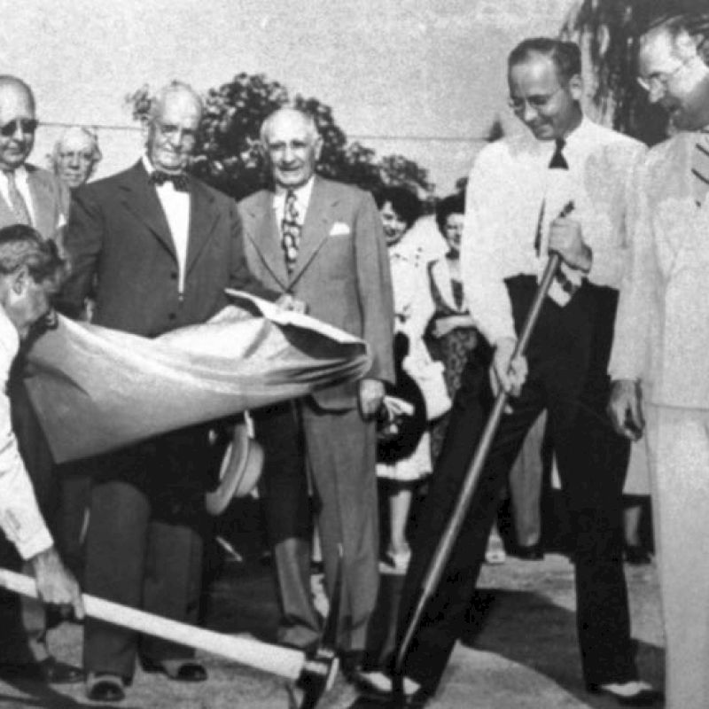 A group of men in formal attire dig and unveil something in a ceremonial setting, with shovels and officials watching, in black and white.