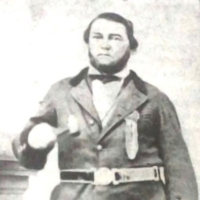 A black‑and‑white photo of a man in old-fashioned formal attire with a belt, standing and gesturing with one hand, indoors.