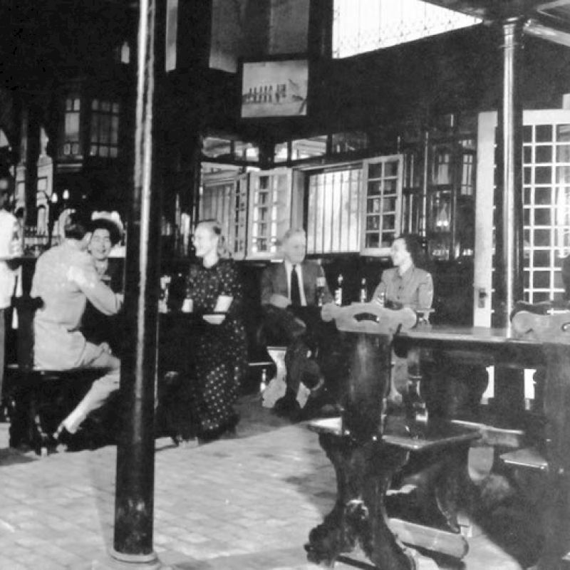 A vintage caf&eacute; scene with patrons seated at long benches and tables, a bartender behind the counter, and a striped pillar in the foreground, time-worn and nostalgic.