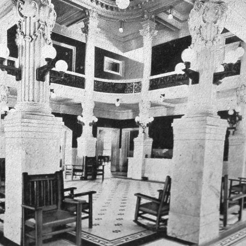 A black-and-white photo of an old temple or hall with pillars, carved columns, wooden benches, and a tiled floor in a circular, balcony-overlook setting, reminiscent of a historic meeting room or temple. Top it at 140 characters, always ending the sentence.