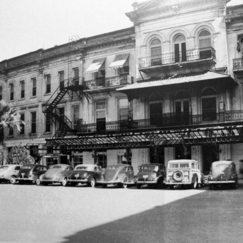 Old black-and-white street scene with a row of vintage cars parked along storefronts; a wrought-iron balcony covers the buildings, era looks mid-20th century.