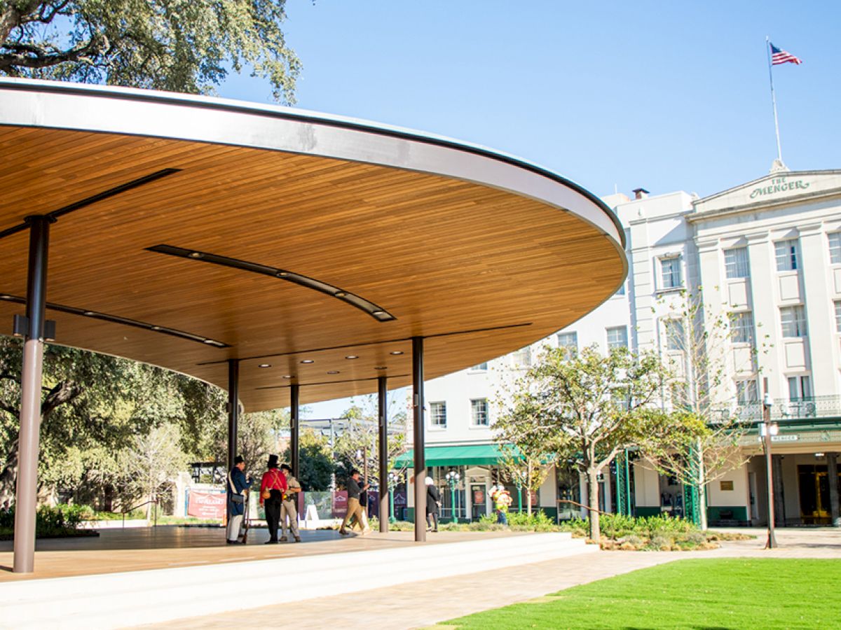 A modern curved pavilion with a wooden underside shades a small group of people outside a white government-style building, sunny day, green lawn.