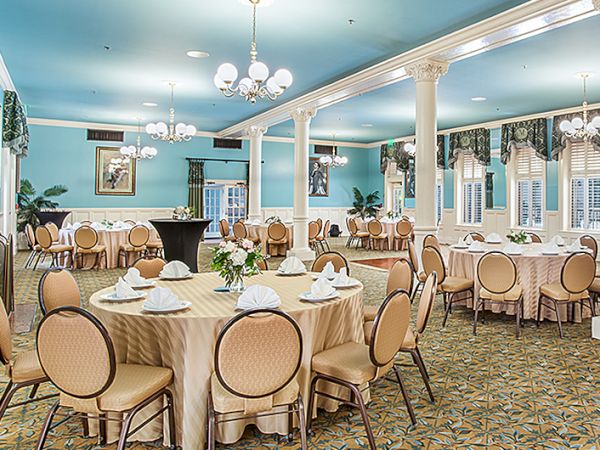A formal banquet hall with round tables, beige chairs, pastel blue walls, chandeliers, and elegant place settings for a refined event.