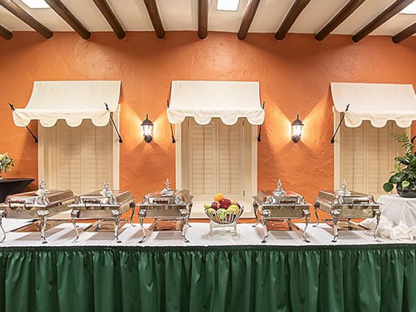 A buffet setup with chafing dishes line a long table against a warm orange wall, three white canopies above, and a fruit centerpiece.