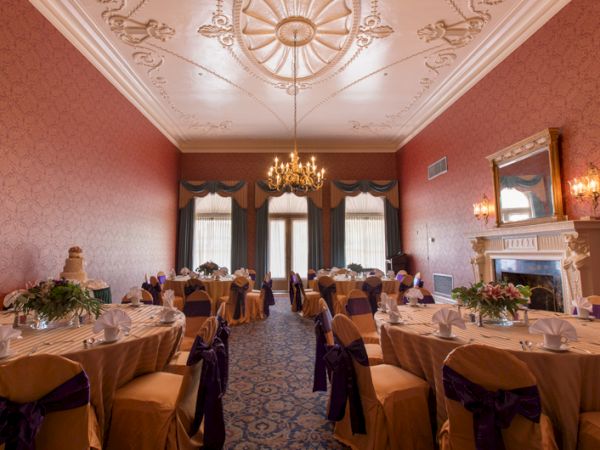 A grand banquet hall with round tables, beige covers and purple sashes, ornate ceiling, chandelier, pink walls, and a classic fireplace &mdash; elegant dining setup.